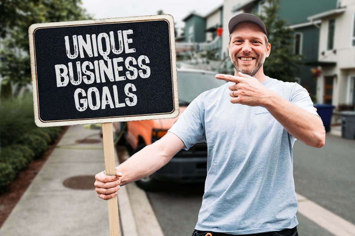 Seattle Window Cleaning owner Martin Skarra standing on a residential street in front of one of his vans holding and pointing to a chalkboard sign that reads "Unique Business Goals"